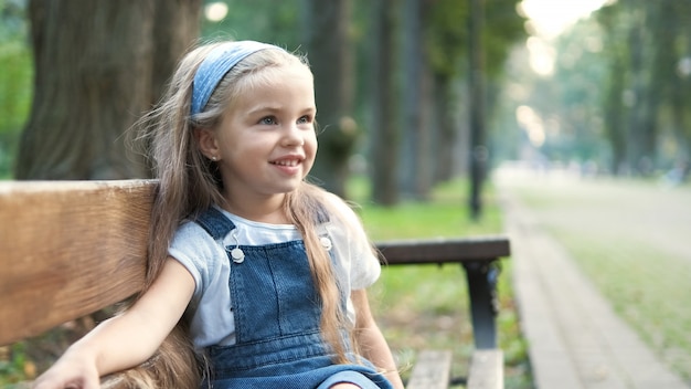 Criança feliz menina sentada num banco sorrindo alegremente no parque de verão.