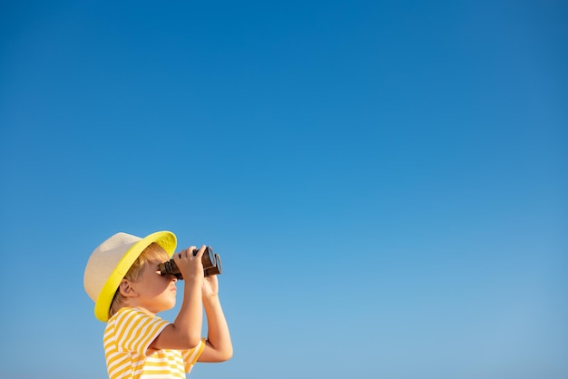 Criança feliz olhando através de binóculos contra o céu azul. Garoto se divertindo ao ar livre no verão. Conceito de aventura e viagem