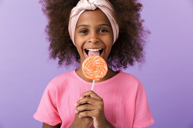 criança feliz otimista jovem africana posando isolado sobre a parede roxa, comer pirulito doce.