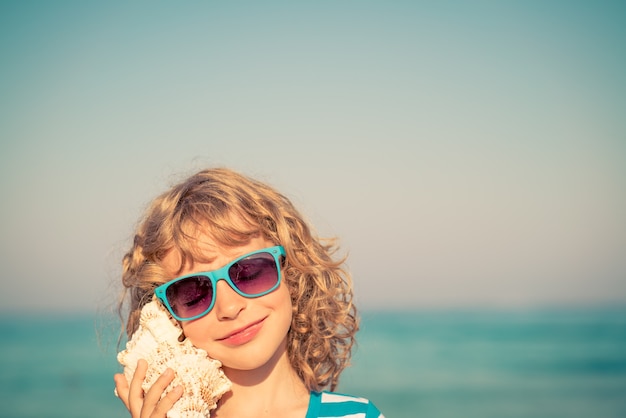 criança feliz ouvindo uma concha na praia contra o fundo do mar e do céu