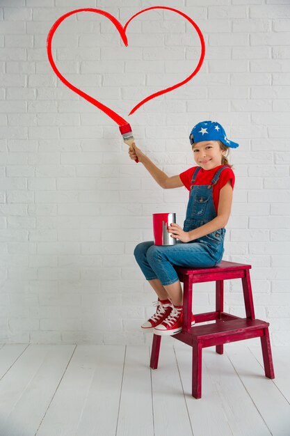 Criança feliz pintando um grande coração vermelho na parede. garota engraçada brincando em casa. cartão de dia dos namorados. renovação e conceito de design