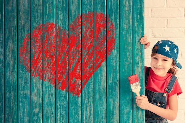 Criança feliz pintando um grande coração vermelho na parede. Garota engraçada brincando em casa. Cartão de dia dos namorados. Renovação e conceito de design