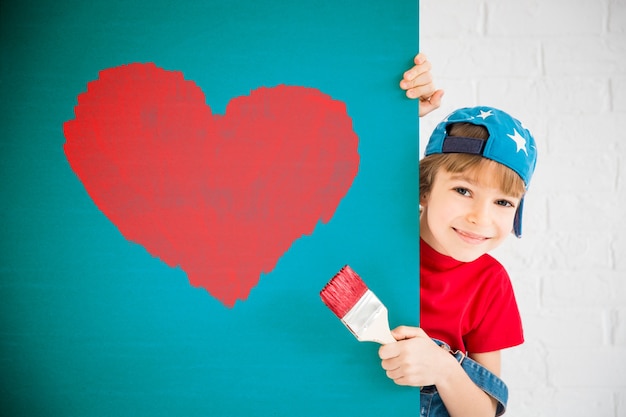 Foto criança feliz pintando um grande coração vermelho na parede. garota engraçada brincando em casa. cartão de dia dos namorados. renovação e conceito de design