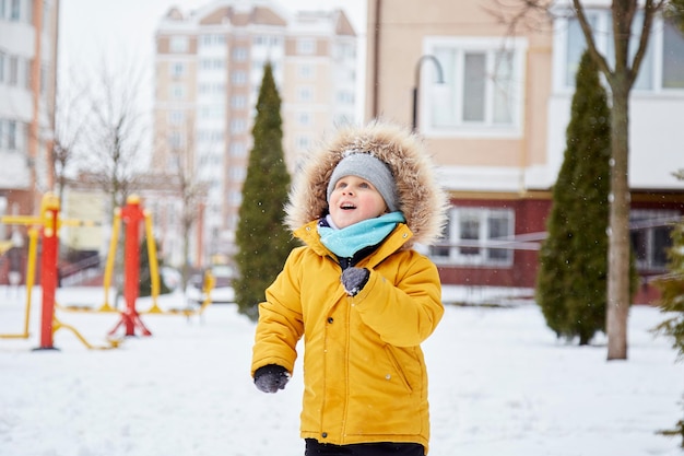 Criança feliz se divertindo na neve na cidade Diversão de inverno lá fora Garoto com jaqueta de inverno laranja brilhante