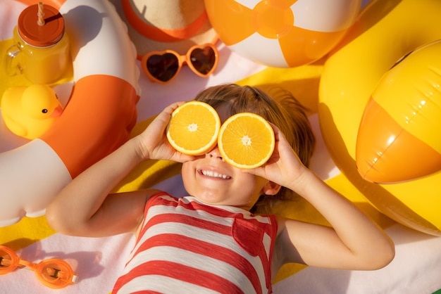 Criança feliz segurando fatias de fruta laranja como óculos de sol Criança vestindo camiseta listrada deitada na toalha de praia Alimentação saudável e conceito de férias de verão