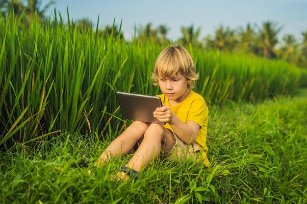 Criança feliz sentada no campo segurando o tablet Menino sentado na grama no dia ensolarado Escola em casa ou jogando um tablet