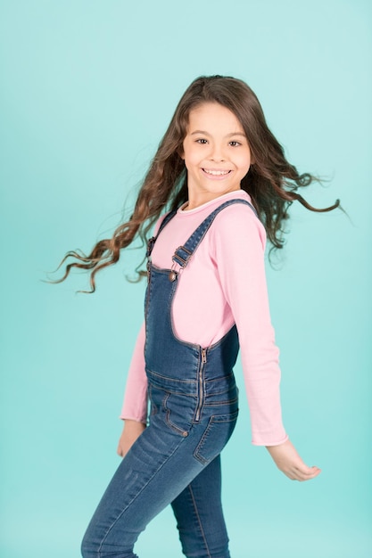 Criança feliz sorrindo com fluindo cabelo longo morena, beleza. Menina pose de jeans em geral, moda. Beleza, olha, penteado. Moda casual. Infância, juventude, tendência pastel vigorosa