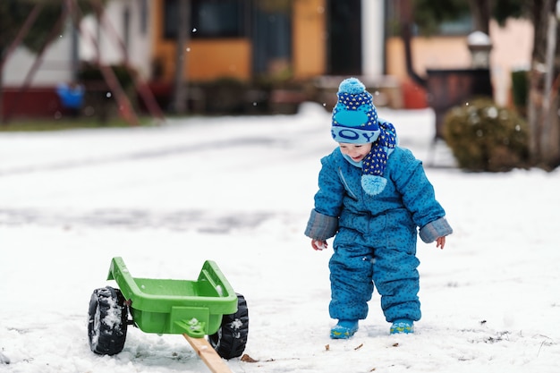 Criança feliz, vestida com roupas quentes, brincando com reboque de brinquedo na neve.