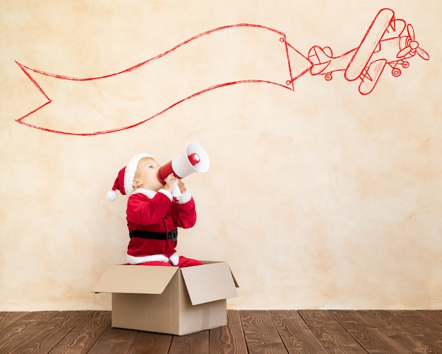 Foto criança feliz vestida de fantasia de papai noel brincando em casa. garoto engraçado dirigindo carro de brinquedo e falando por megafone. conceito de férias de natal