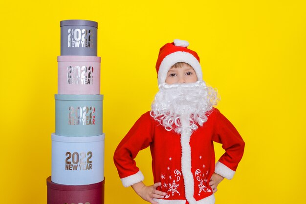 Criança fica ao lado de uma torre de caixas redondas com ano novo 2022, ela está vestida com fantasia de papai noel