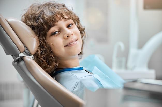 Criança fofa de olhos castanhos sorrindo enquanto está sentada em uma cadeira odontológica, pronta para fazer um check-up dentário