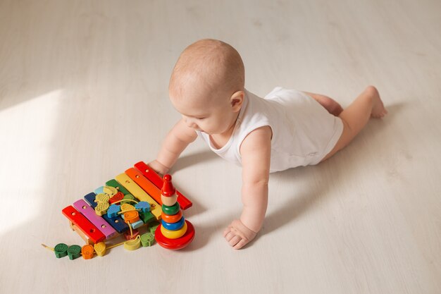 Criança fofa em um macacão branco deitada no chão de madeira da casa com um brinquedo em desenvolvimento