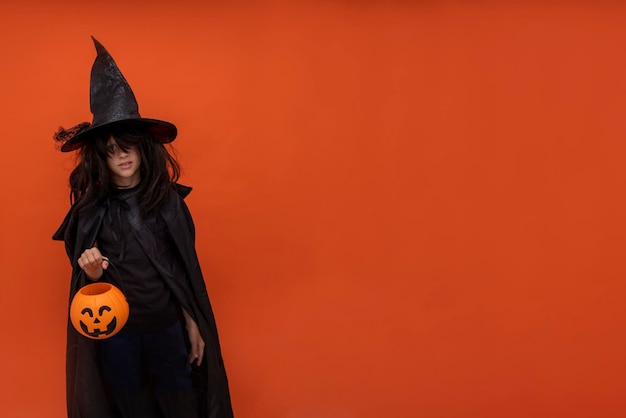 Criança halloween Um menino em uma fantasia de feiticeiro em um fundo laranja Menina em uma fantasia de bruxa Copiar espaço Jack o lanterna Doces em uma abóbora Lugar para texto Terno preto escuro