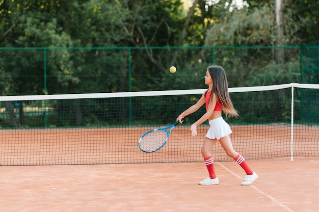 Criança jogando tênis na quadra ao ar livre. Menina com raquete de tênis e bola no clube de esporte. Exercício ativo para crianças