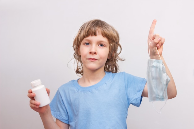 Criança loira de camiseta azul, segurando a máscara médica e frasco branco com remédio nas mãos. Prevenção da propagação da infecção por vírus e conceito de contaminação