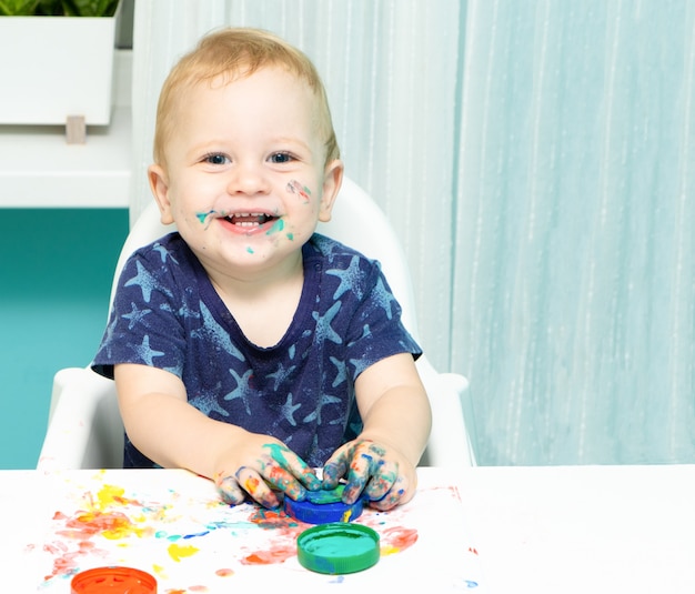 Criança loira sorrindo, desenhando com tintas de um dedo