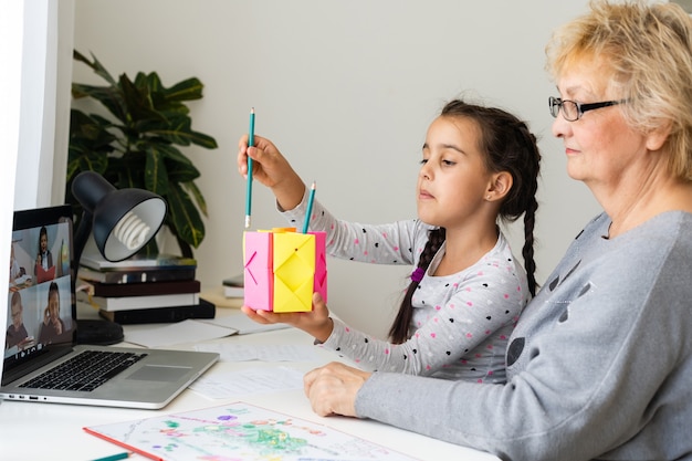 Criança menina bonita e feliz usando o computador portátil com a avó, estudando através do sistema de e-learning online.