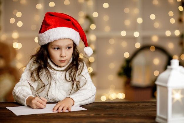 Criança menina feliz com chapéu de Papai Noel escrevendo uma lista de desejos ou uma carta para o Papai Noel