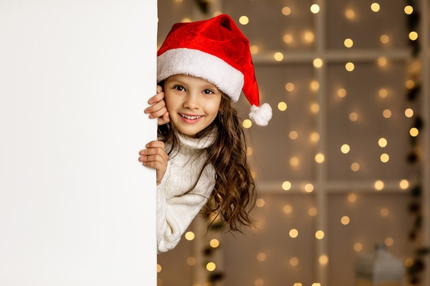 Criança menina feliz com chapéu de Papai Noel vermelho segurando banner de papelão branco em branco com espaço vazio