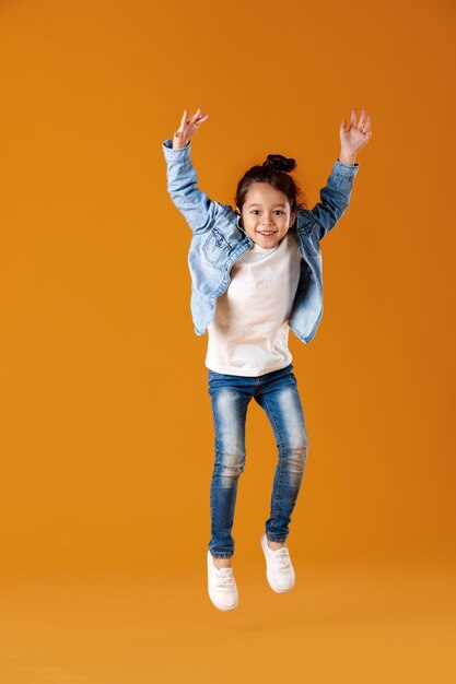 Criança menina feliz com roupas jeans pulando em um fundo laranja