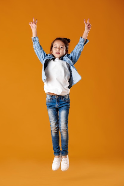 Criança menina feliz com roupas jeans pulando em um fundo laranja