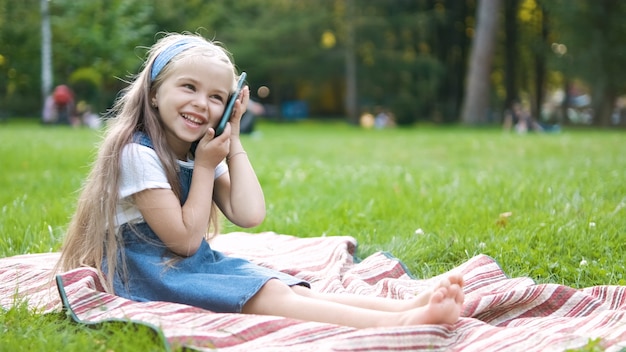 Criança menina feliz tendo uma conversa falando em seu sellphone no parque de verão.