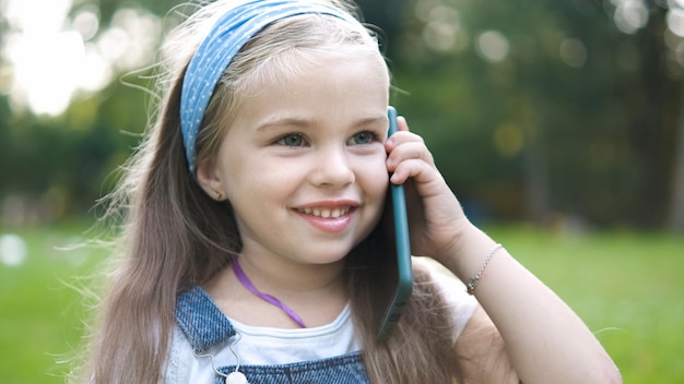 Criança menina feliz tendo uma conversa falando em seu sellphone no parque de verão.