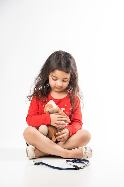 Criança menina indiana com estetoscópio e brinquedo de bebê ou filhote de cachorro de pelúcia sentada contra um fundo branco