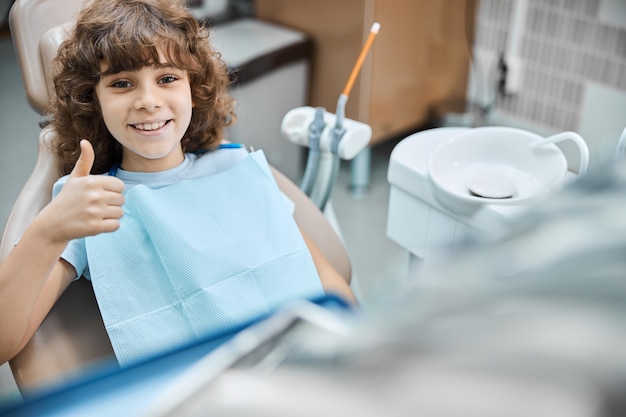 Foto criança morena encantadora sorrindo e segurando o polegar enquanto espera para o exame odontológico