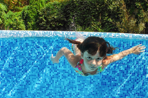 Criança nada na piscina debaixo d'água, feliz garota ativa mergulha e se diverte na água, criança fitness e esporte em férias em família