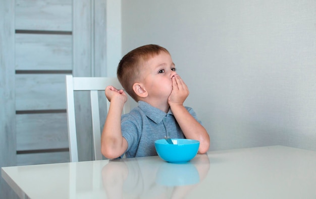 Criança não quer comer e empurra o prato, um garotinho está na cozinha. Uma criança triste não quer comer mingau. Um menino come sem apetite. TConcepto de pequeno-almoço saudável para crianças.