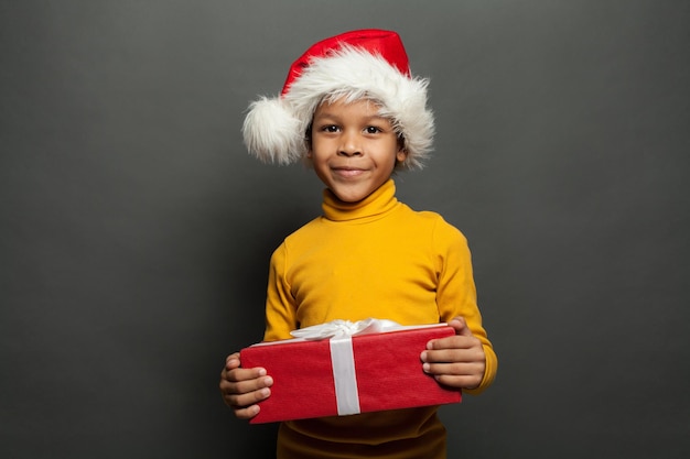 Criança negra bonita com chapéu de Papai Noel segurando um presente de Natal vermelho em fundo cinza escuro