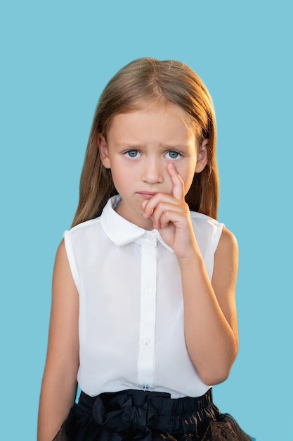 Criança ofendida punição injusta triste decepção retrato de menina doce chorando infeliz com roupa elegante, limpando a lágrima com o dedo isolado no fundo azul