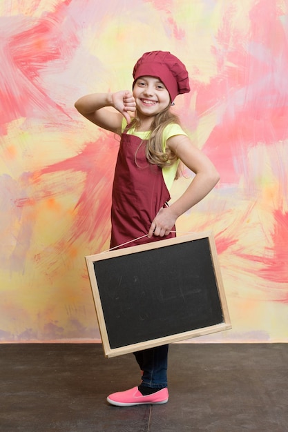 Criança ou garotinha feliz cozinha com chapéu de chef vermelho e avental segura lousa de menu em fundo colorido cozinhando e comendo espaço de cópia