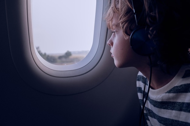 Criança ouvindo música em fones de ouvido e olhando pela janela do avião escuro durante o voo à noite