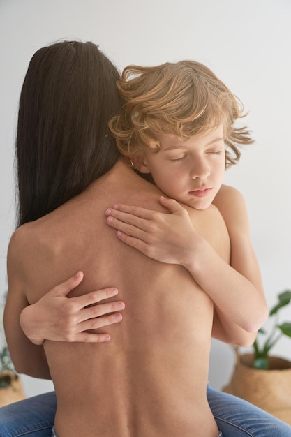 Criança pacífica, cabelos cacheados e olhos fechados, abraçando ternamente a mãe anônima em topless enquanto está sentada em uma sala iluminada em casa
