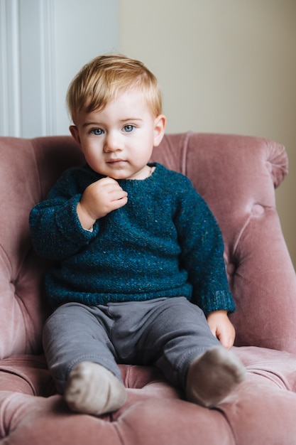 Criança pequena bonita com bochechas gordas, cabelo loiro, veste roupas da moda, senta-se na poltrona