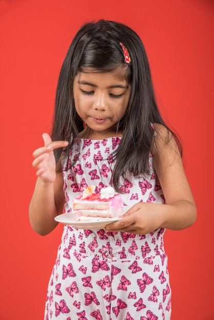 Criança pequena bonita indiana ou asiática comendo um pedaço de massa ou bolo com sabor de morango ou chocolate em um prato. Isolado sobre fundo colorido