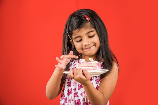 Criança pequena bonita indiana ou asiática comendo um pedaço de massa ou bolo com sabor de morango ou chocolate em um prato. Isolado sobre fundo colorido
