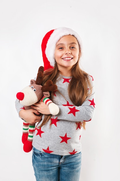 Criança pequena bonitinha no chapéu de Papai Noel com cervo de brinquedo de malha. A menina rindo e apreciando o presente. Conceito de natal