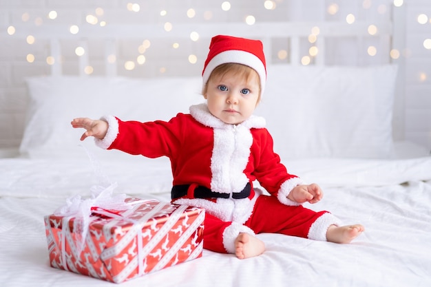 Criança pequena com fantasia de Papai Noel segurando um presente de Natal sentada em um fundo branco de ano novo