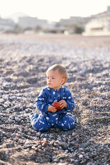 Criança pequena em um macacão azul sentada de joelhos em uma praia de seixos e segurando uma maçã close-up