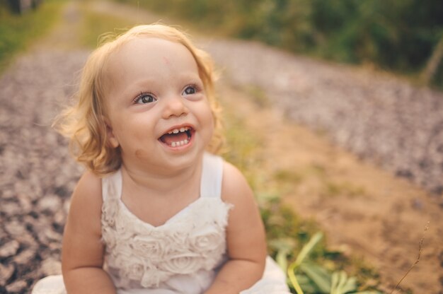 Criança pequena engraçada loira bonita criança com cachos em um vestido branco e com lama no rosto senta-se