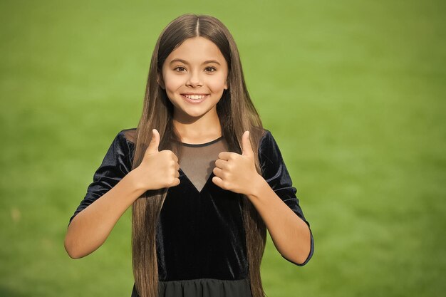 Criança pequena feliz com cabelo comprido e aparência de beleza usa vestido de moda mostrando polegares para cima gesto de mãos grama verde verão aprovação ao ar livre