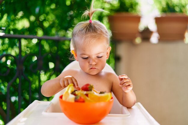 Criança pequena sentada em uma cadeira alta na varanda e olhando para um prato de frutas