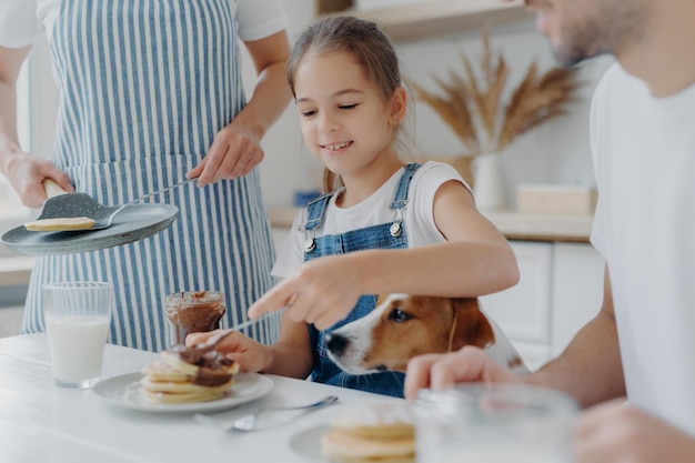 Criança pequena, seu pai e cachorro sentam-se juntos na mesa da cozinha, comem panquecas recém-preparadas, mãe de avental fica perto da panela. Família apetitosa sobremesa saborosa na cozinha. Cozinhar, conceito de nutrição