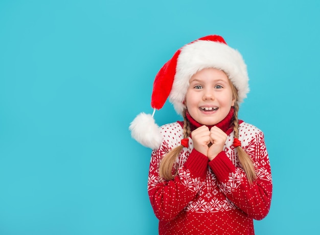 Criança saiu no suéter de Natal de chapéu de Papai Noel esperando por Papai Noel ou presentes em azul com espaço de cópia