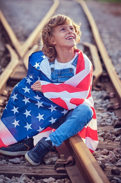 Criança satisfeita envolta na bandeira dos EUA e olhando para cima enquanto está sentada na ferrovia durante a celebração do Dia da Independência