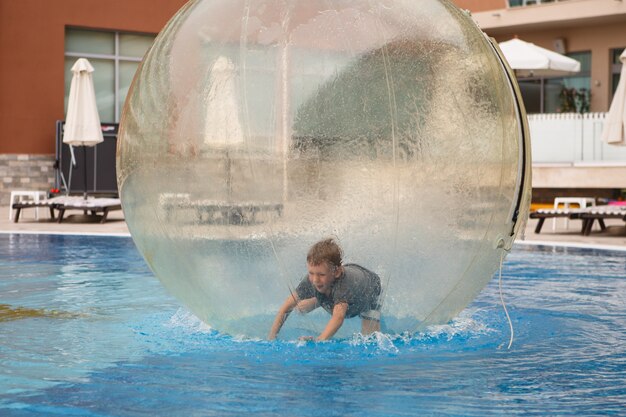 Foto criança se diverte dentro de um grande balão de plástico na água da piscina do resort de verão. menino dentro de uma grande bola inflável transparente correndo e se divertindo.