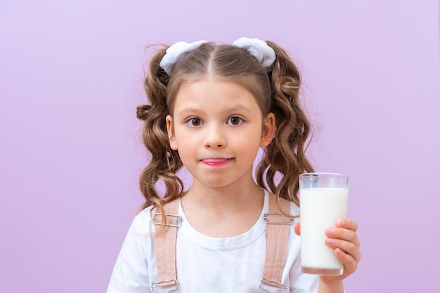 Criança segura um copo de leite e lambe os lábios. bigode de espuma de leite. Uma menina bebe leite no café da manhã.